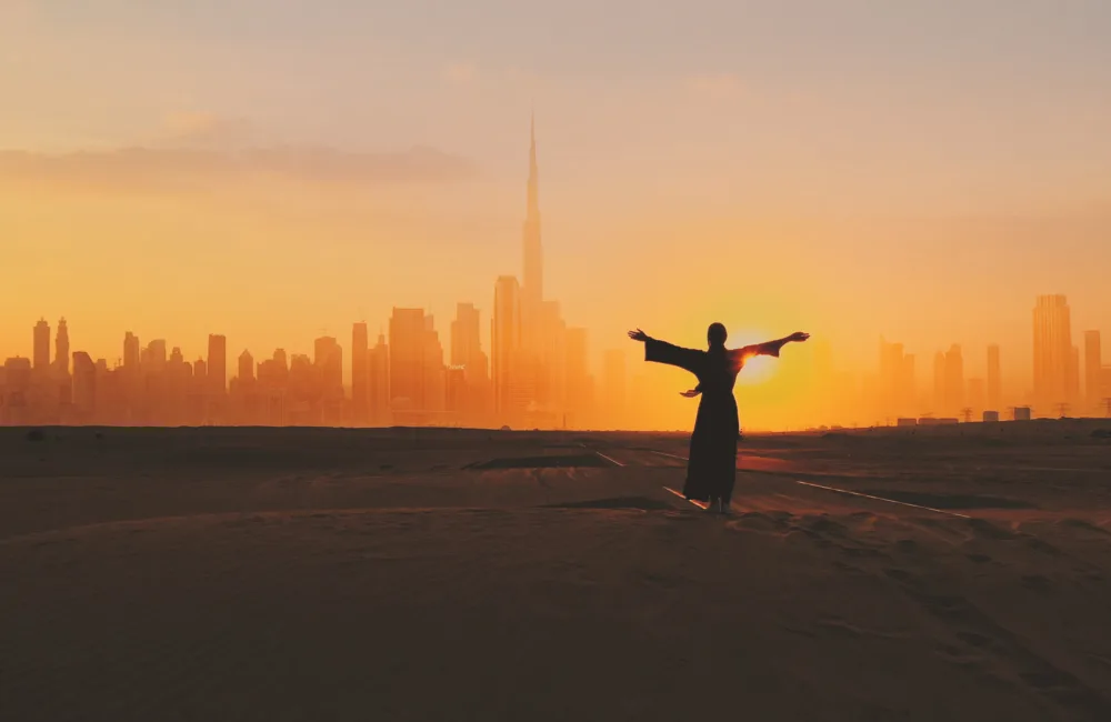 Emirati woman wearing the traditional UAE dress while rising her hands on the sunset at a desert with the silhouette of downtown Dubai on the background.