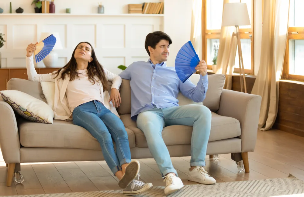 Exhausted man and woman feeling discomfort in the living room while waving blue paper fans due to the hot climate.