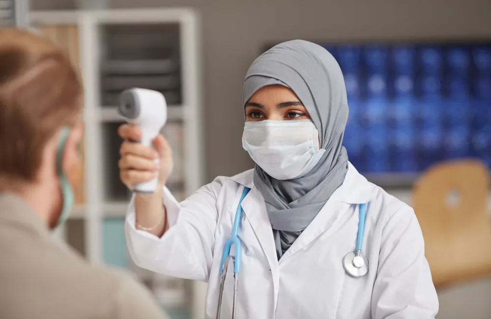 A female doctor wearing a hijab taking the temperature of a patient using a digital thermometer.