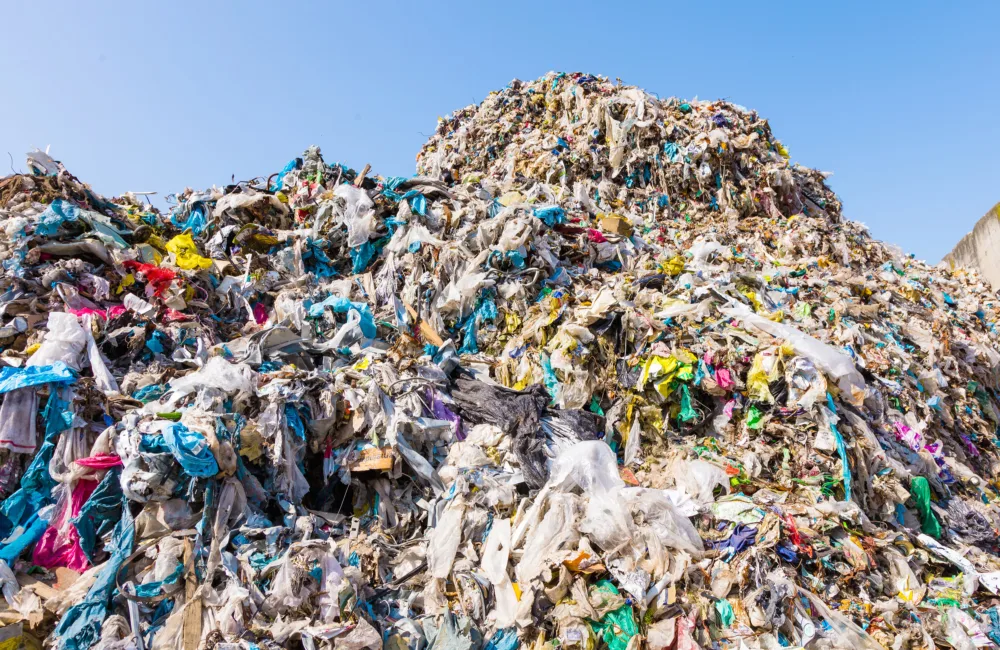 A large, densely packed pile of mixed waste, mainly plastic bags in various colors, sits outdoors beneath a clear blue sky. No people or text are visible in the image.
