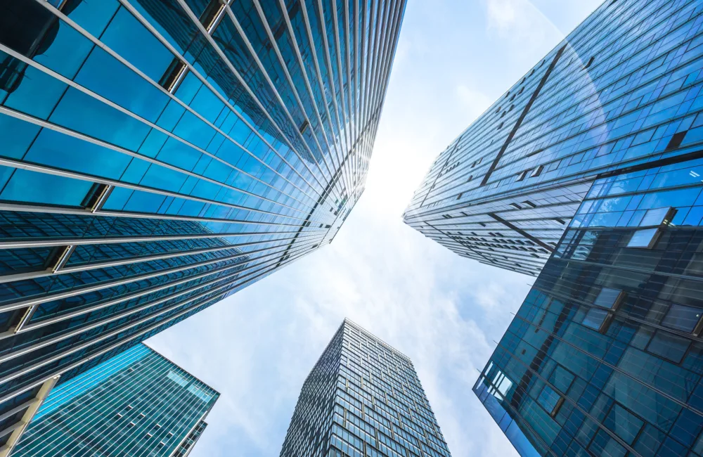 A low‑angle view looking upward at several tall glass‑clad skyscrapers reflecting the sky. The buildings form a circular frame around a bright, cloud‑dappled sky at the centre.