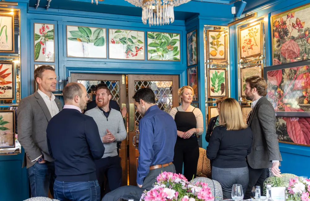 Group of adults standing and talking inside a brightly decorated room with teal walls, framed botanical illustrations, a chandelier, and double doors; table in foreground set with flowers, glassware, and bottles.