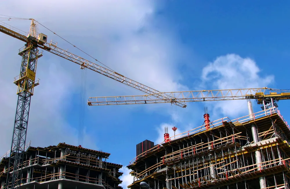 Two cranes lifting building materials to two partially constructed multi-storey buildings against a blue sky