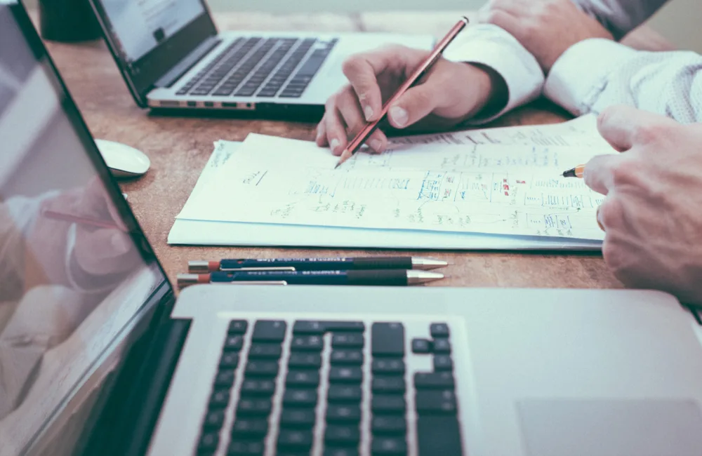 Close up of the hands of two people sitting at a desk, planning on paper and laptops