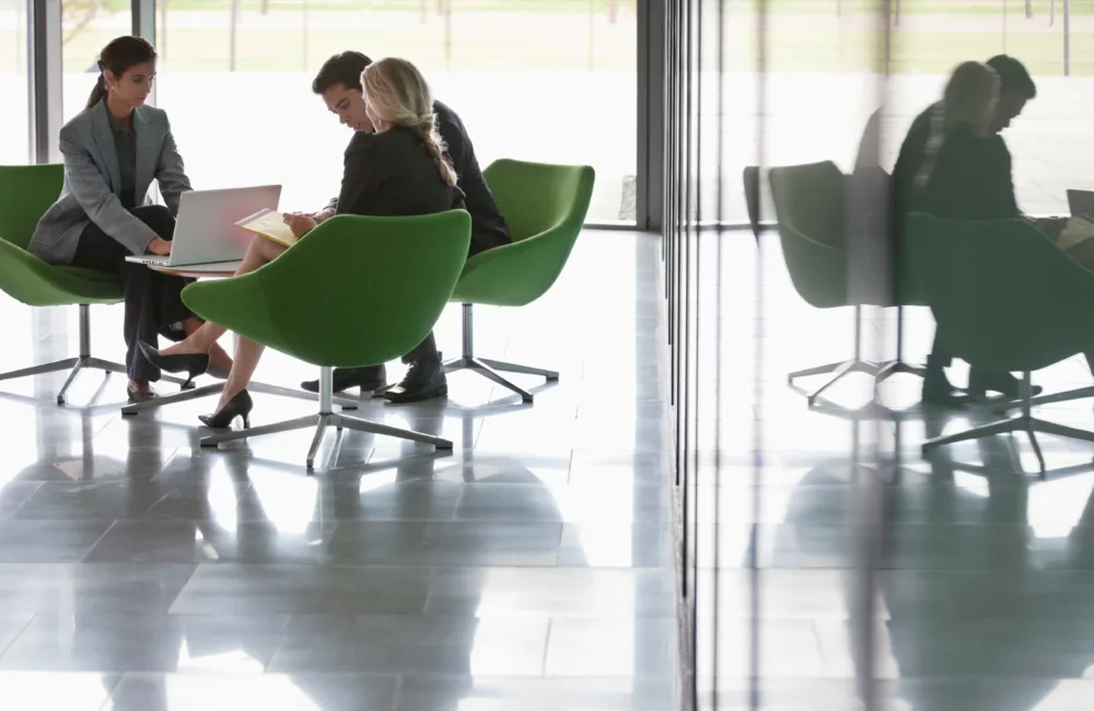 Group of 3 smartly dressed people sitting in green chairs in an office space discussing something on a laptop