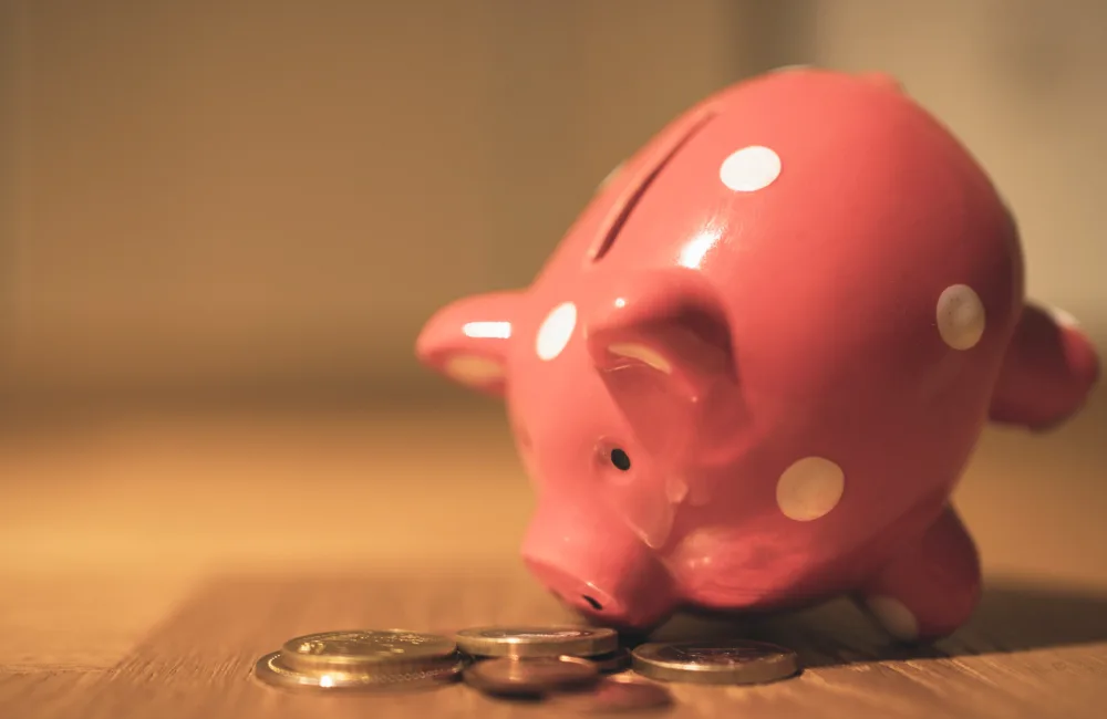 A pink piggy bank with white spots leaning over nose first onto a small pile of coins
