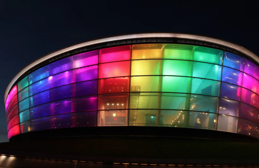 Glasgow SEC centre (a large round arena) viewed from the outside, internally lit up in rainbow colours against a deep blue night sky