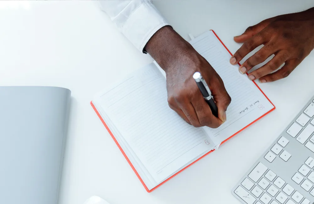 Overhead view of a person's hands writing in a white and red notebook on top of a white desk. On the left is a grey notebook and the right is a thin grey and white keyboard.