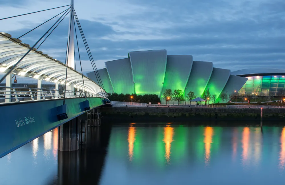 The Glasgow SEC centre and Armadillo buildings from across the River Clyde. The Bell Bridge is visible to the left, the buildings are lit by coloured lights.