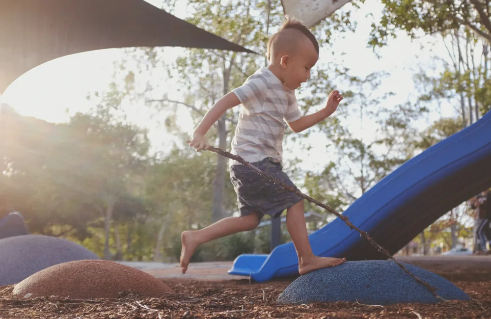 Child running barefoot in front of a slide on a soft bark floor in an outdoor play area
