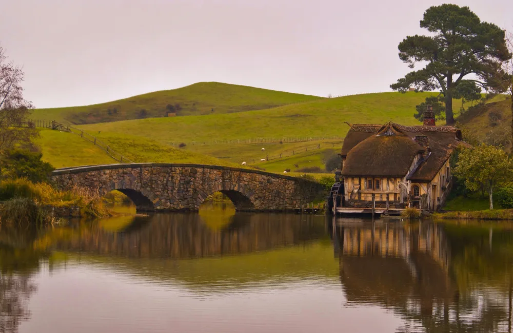 Old sone bridge with two arches over a still lake leading to an old thatched yellow house