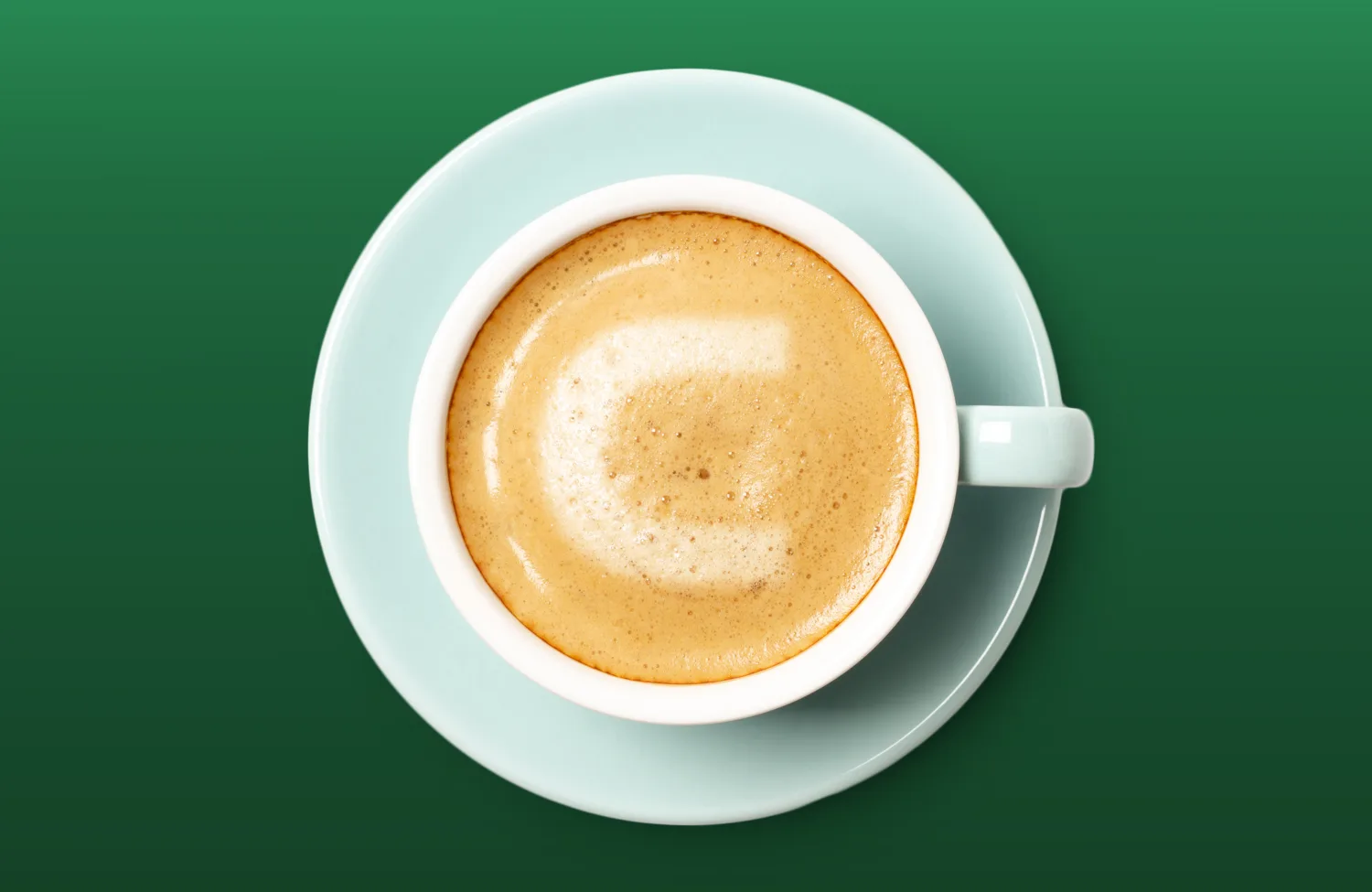 Top-down view of a cup of frothy coffee in a light mint‑green cup and saucer, placed against a smooth green background. The foam forms a subtle C shape on the surface.