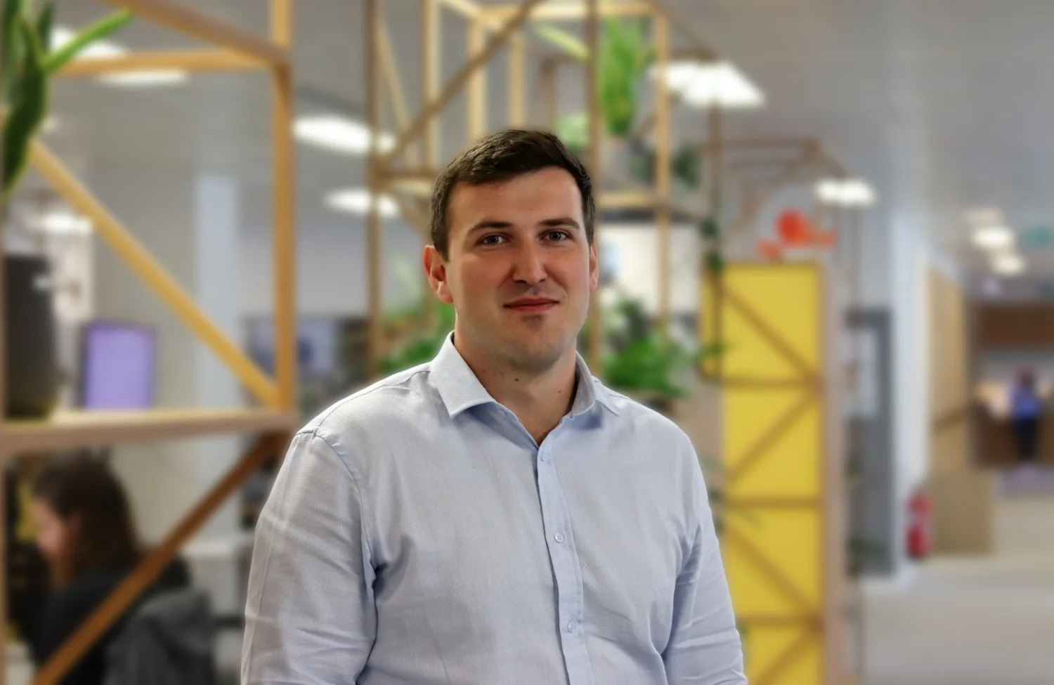 Person standing indoors in a modern office, wearing a light blue button-down shirt. Wooden shelving with plants and a yellow partition appear behind, with desks and other people in the background.