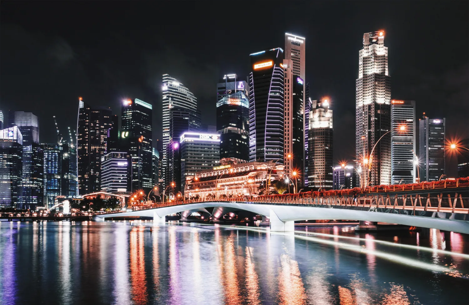 Night time shot of singapore including high rise buildings a bridge over the river and the merlion on the left hand side