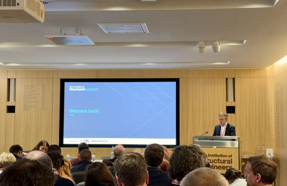 A conference room with attendees seated facing a large screen displaying the text ‘Welcome back!’ and the logo of the Institution of Structural Engineers. A person stands at a podium labelled with the same institution name.