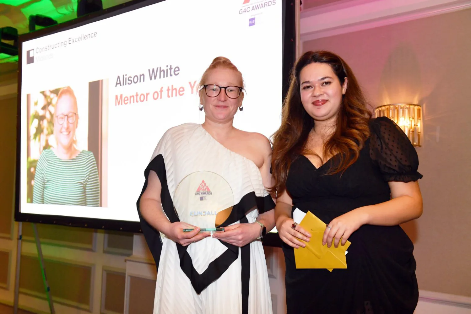 Alison on stage in a long white dress holding her award  with the host in front of the projection screen