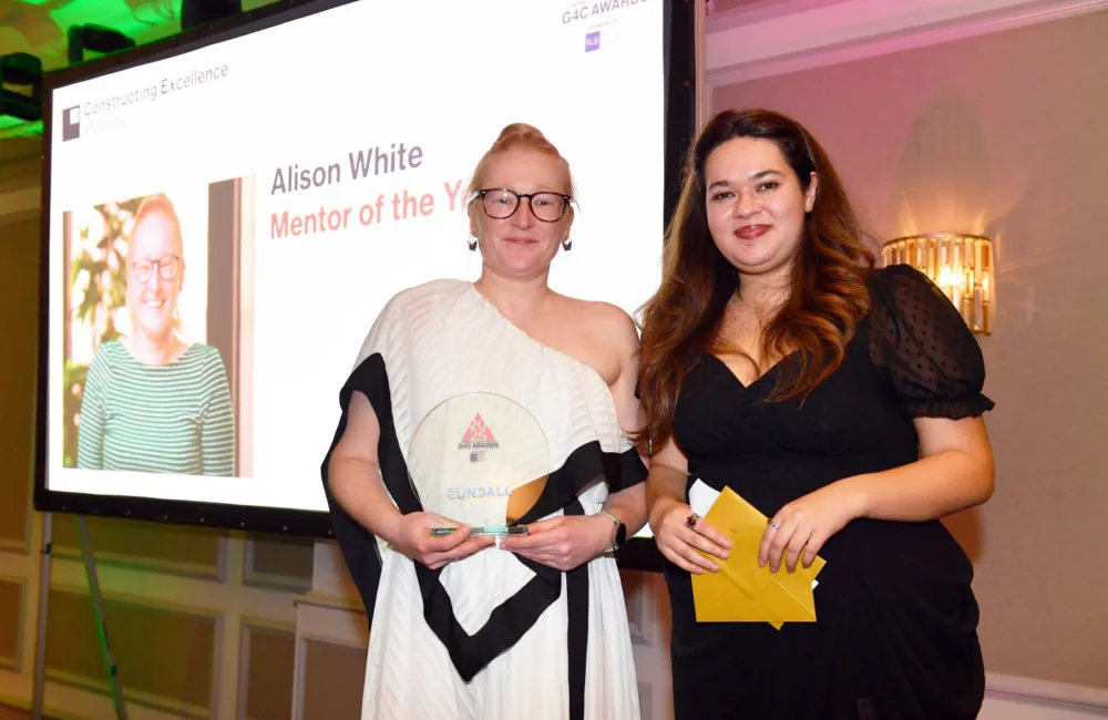 Alison on stage in a long white dress holding her award  with the host in front of the projection screen