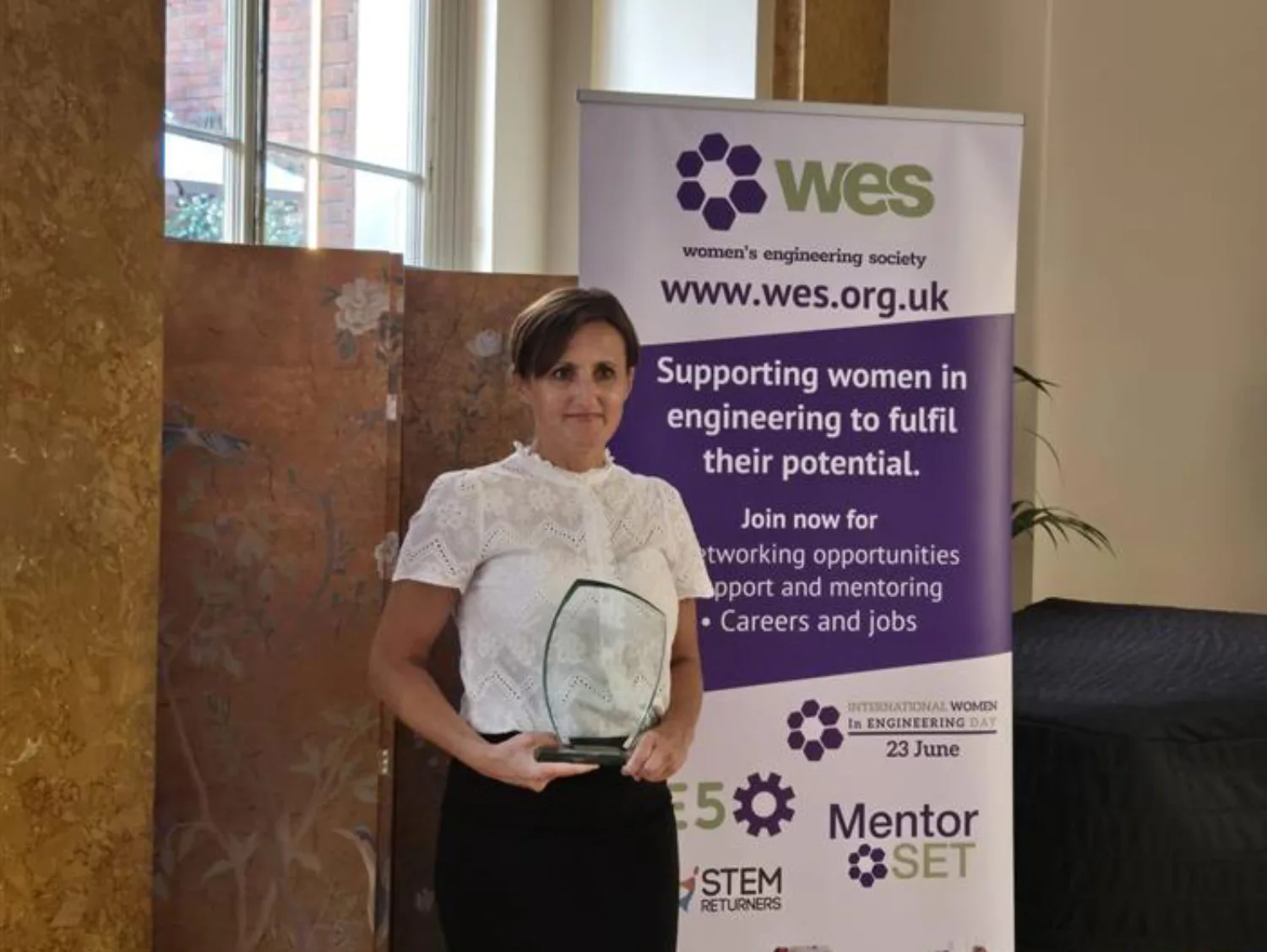 Person holding a glass award stands in front of a Women's Engineering Society banner promoting support, mentoring, and careers for women in engineering. Logos for WES, Mentor SET, and International Women in Engineering Day are visible.