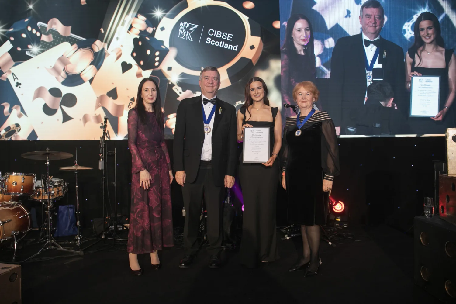 Four people standing on a stage in formal attire, two wearing medals. One person holds a framed certificate. Behind them is a large screen displaying ‘CIBSE Scotland’ with playing card graphics. A drum set is visible on the left.