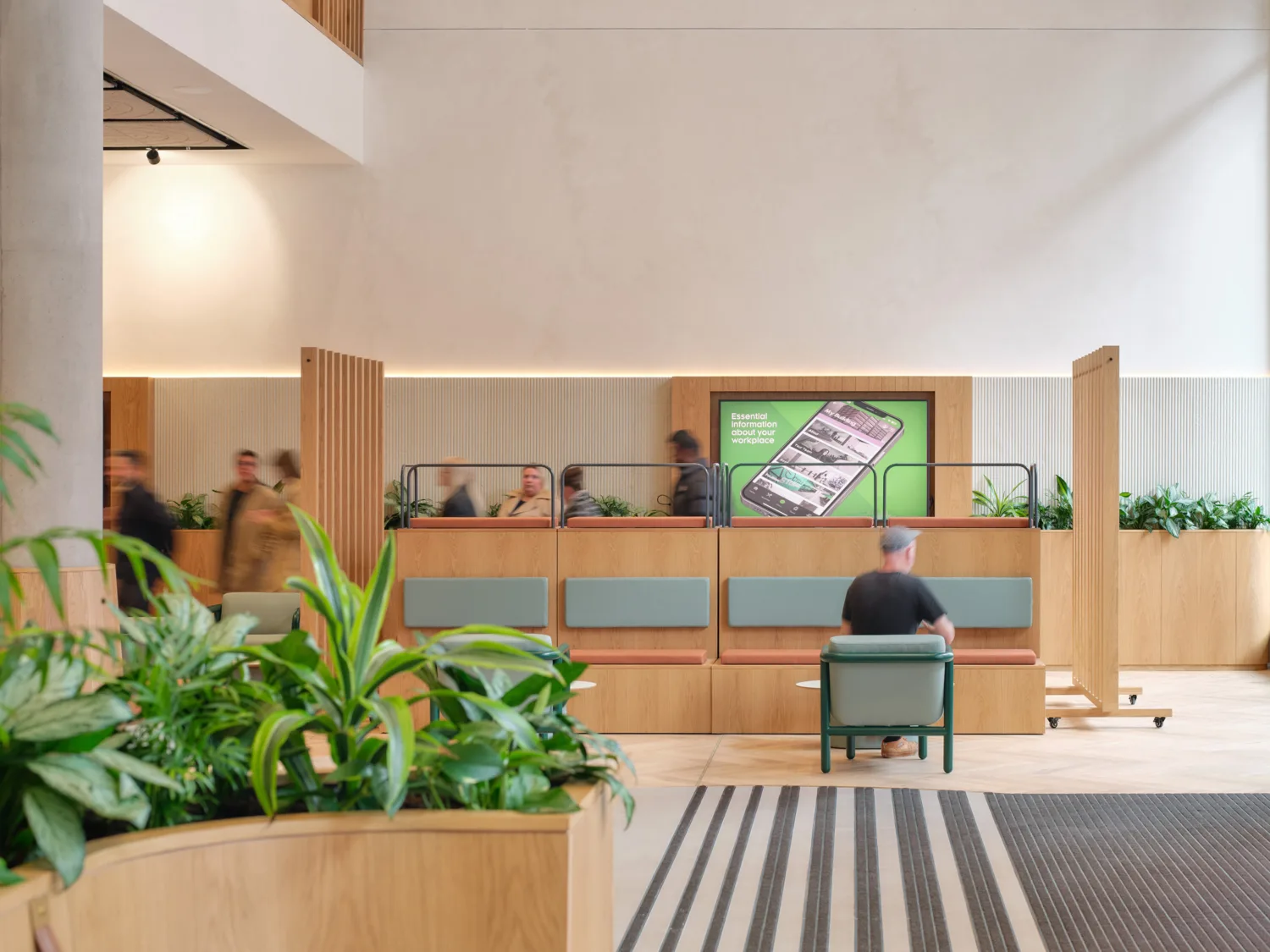 Interior of a modern lobby with light wood partitions, built-in bench seating, and potted plants. Several people walk past while one person sits on a chair. A wall display shows a smartphone graphic with text about workplace information.