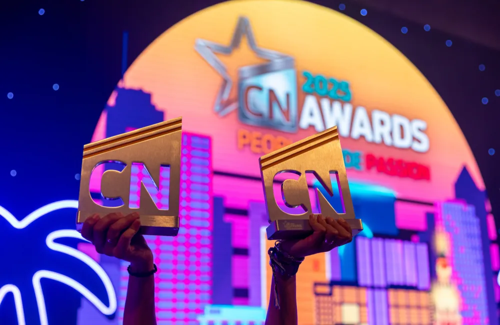 Two hands holding golden trophies engraved with 'CN' in front of a colourful backdrop showing a cityscape and the text '2023 CN AWARDS PEOPLE DE PASION' with a star logo.