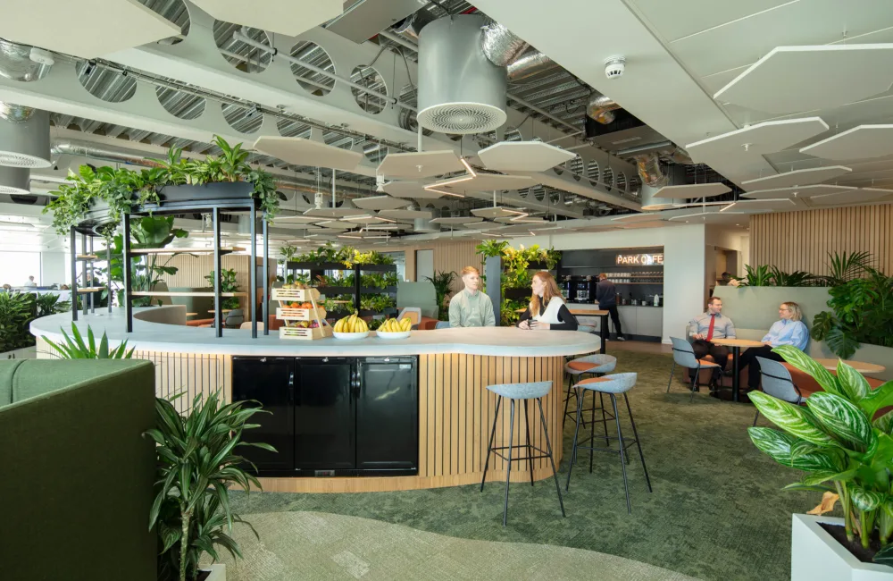 Modern office space with a curved white counter featuring stools, fruit bowls, and a display stand. Surrounding the counter are green plants, wooden slatted panels, and seating areas with tables and chairs. The ceiling has exposed ducts, circular vents, and suspended acoustic panels.