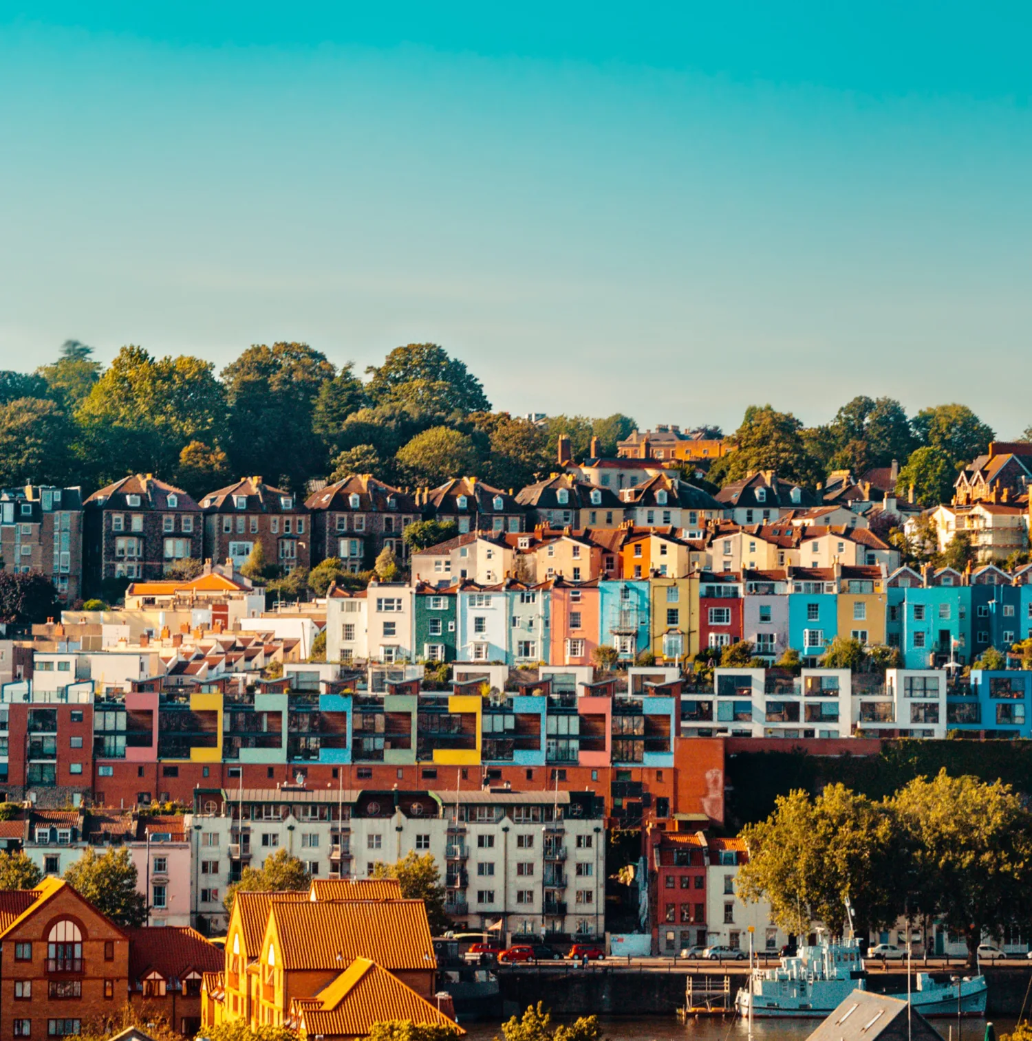 A hillside of colourful Bristol houses behind the canal with a row of trees and blue sky
