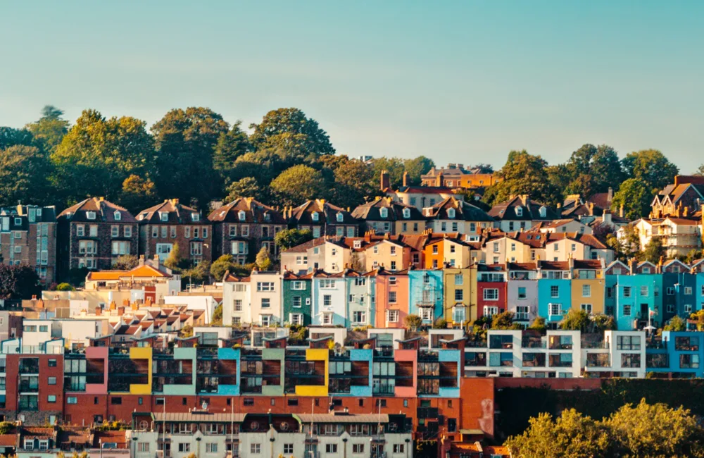 A hillside of colourful Bristol houses behind the canal with a row of trees and blue sky