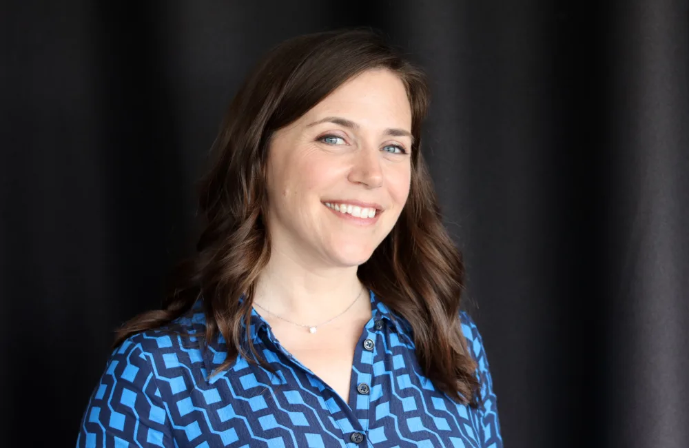 Frances in a light and navy blue top against a dark background smiling to camera