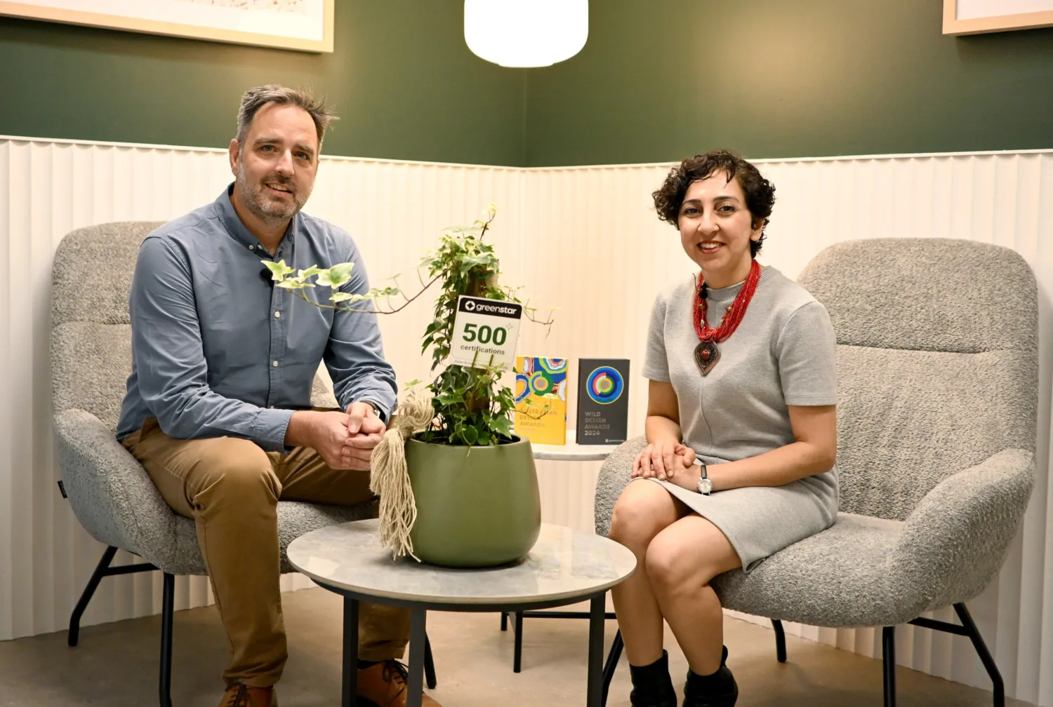 Two people seated in grey upholstered chairs around a small round table with a green potted plant. Behind them is a green and white wall with framed artwork. On the table are items including a sign reading ‘Green Star 500’ and two books.