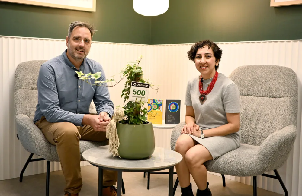 Two people seated in grey upholstered chairs around a small round table with a green potted plant. Behind them is a green and white wall with framed artwork. On the table are items including a sign reading ‘Green Star 500’ and two books.