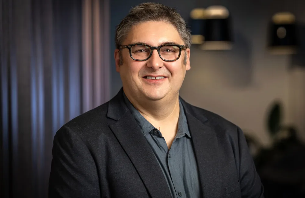 Stewart Mann wearing a dark blazer over a buttoned shirt stands indoors against a softly lit background with dark curtains and hanging light fixtures