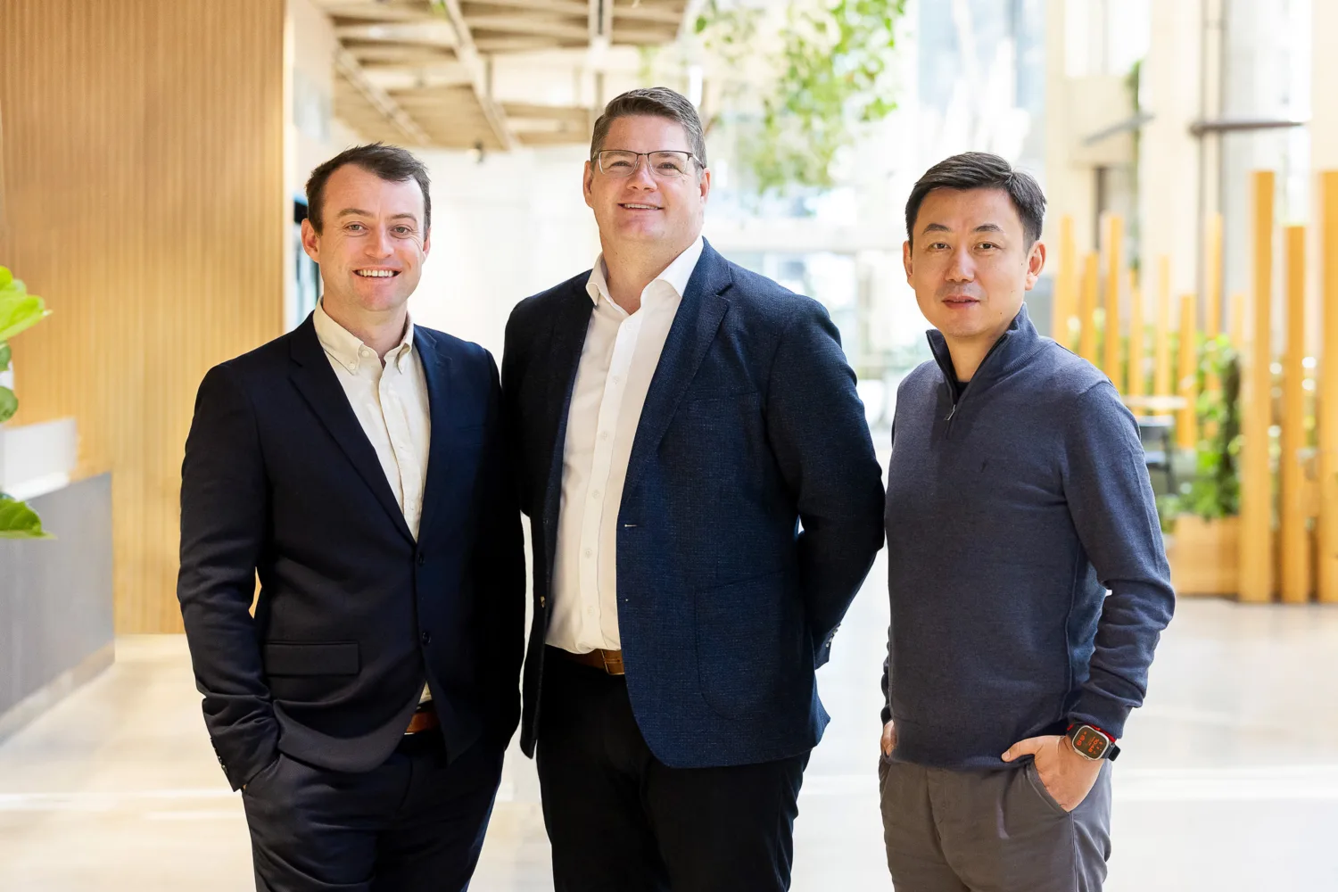 Three individuals stand indoors in a modern office space with wooden decor and plants. One wears a dark suit, another a dark blazer, and the third a casual sweater.