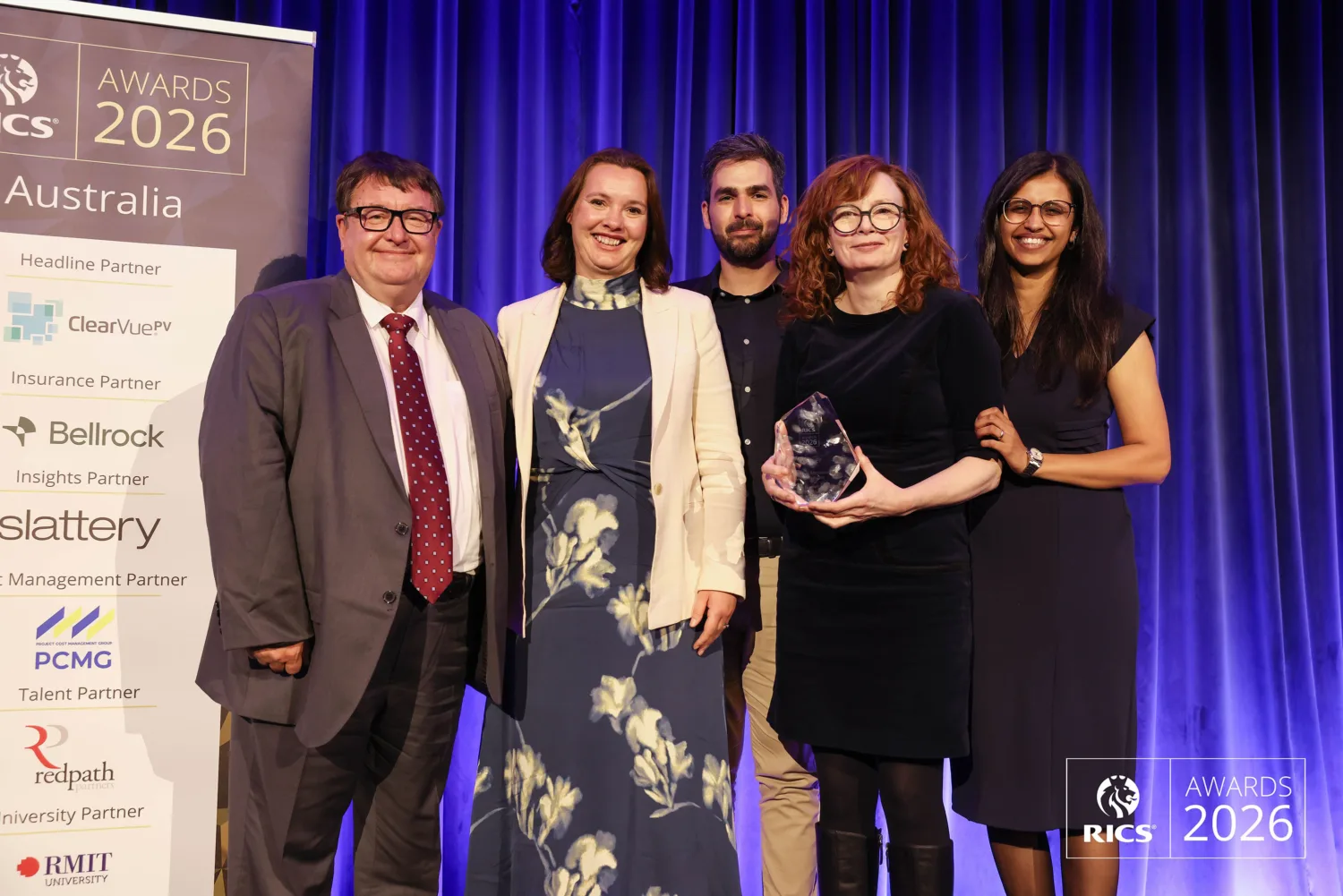 Five people posing on stage at the RICS Awards 2026 Australia event, with one person holding a glass award in front of a blue curtain backdrop.