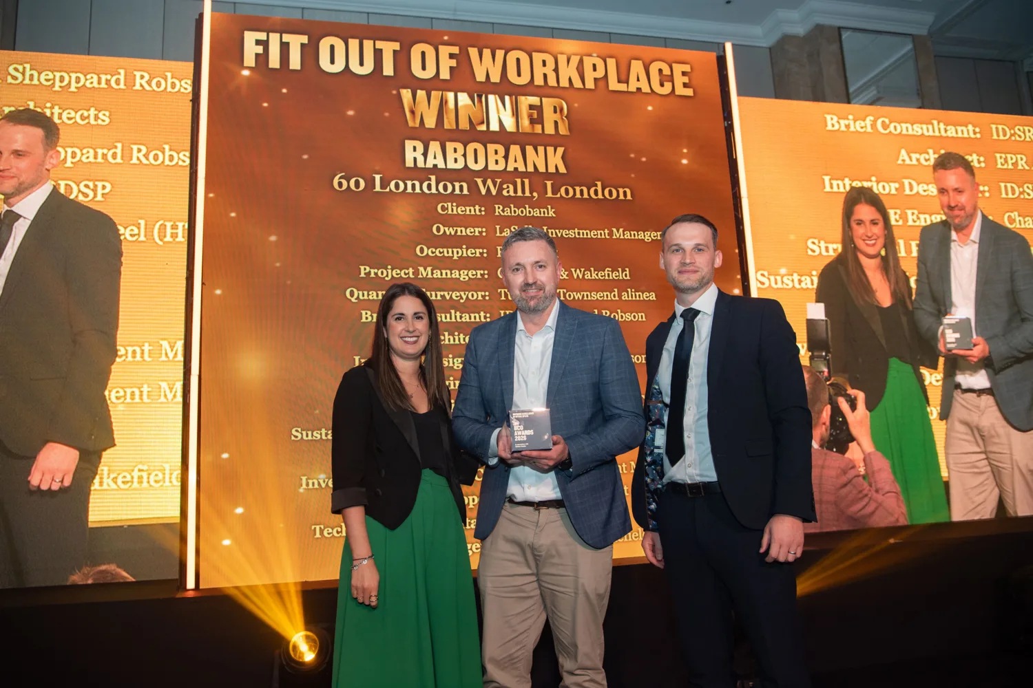Awards stage with a gold backdrop reading “Fit Out of Workplace Winner – Rabobank, 60 London Wall, London,” followed by project details. Three people stand at center holding a glass award, with presentation screens and stage lighting around them.