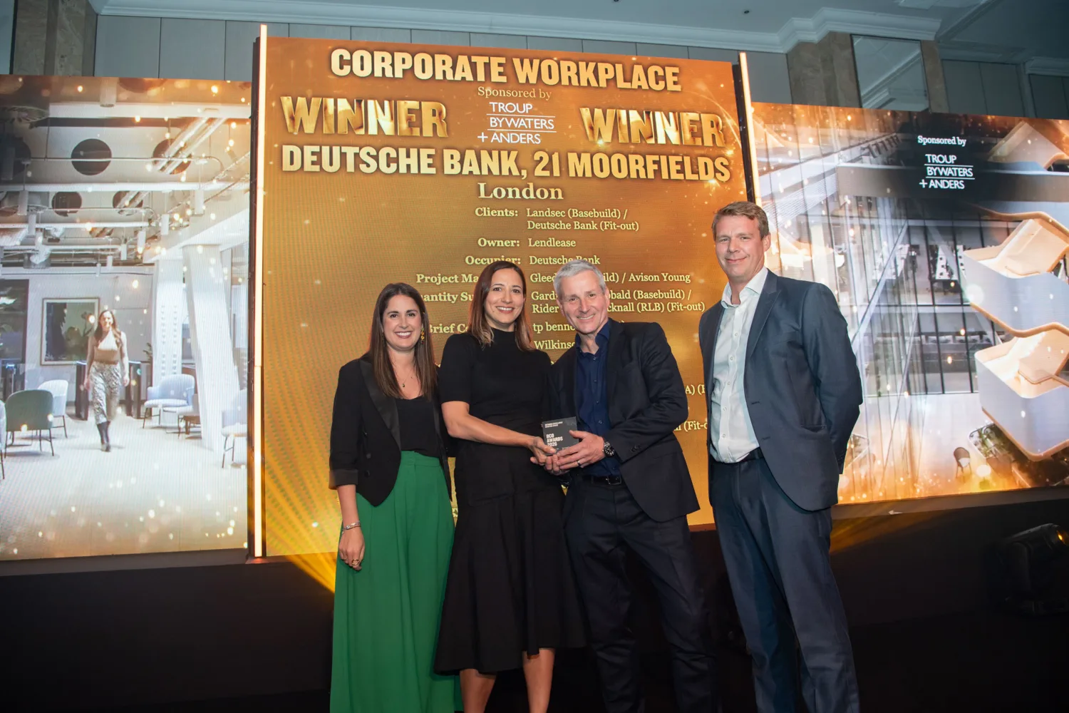 Awards stage with a gold backdrop reading “Corporate Workplace Winner – Deutsche Bank, 21 Moorfields, London.” Four people stand in front holding a glass award, with additional project details listed on the display behind them.