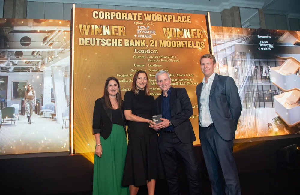 Awards stage with a gold backdrop reading “Corporate Workplace Winner – Deutsche Bank, 21 Moorfields, London.” Four people stand in front holding a glass award, with additional project details listed on the display behind them.