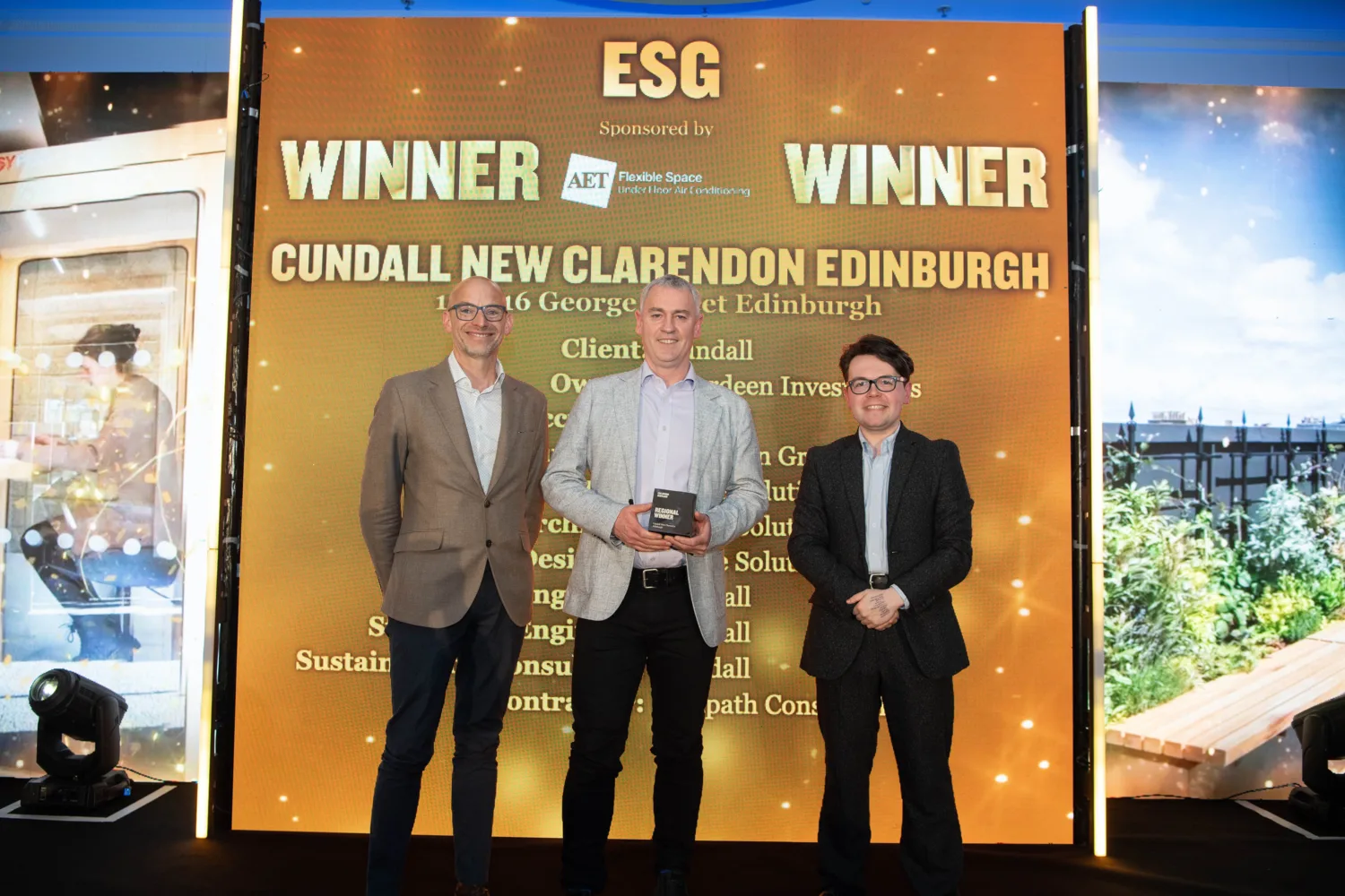 Three people stand on a stage holding an award in front of a large screen reading “ESG Winner.” The backdrop text includes “Cundall New Clarendon Edinburgh,” sponsor AET Flexible Space, and additional project details, with event lighting around the stage.