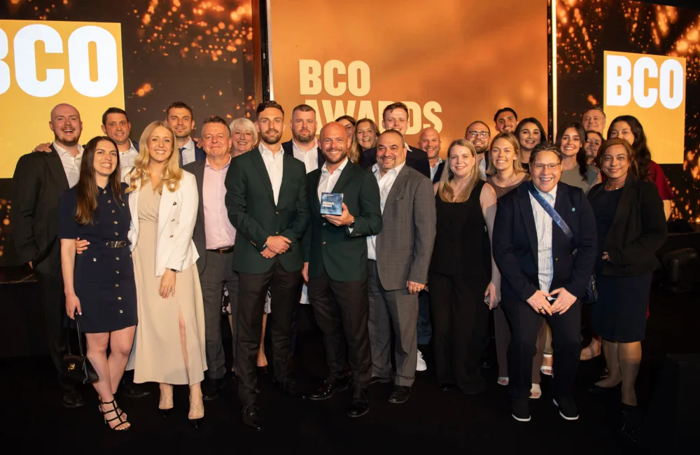 A crowd of people stood on an orange lit stage in front of BCO awards logo signage. The central person is holding the award.