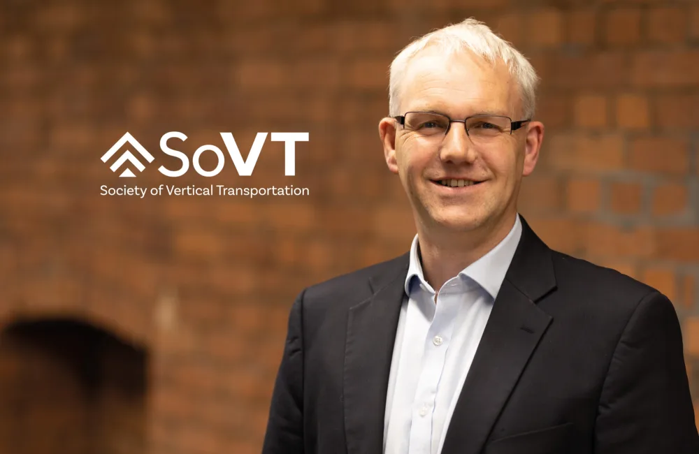 Portrait of a person wearing glasses, a dark blazer, and a light button-up shirt, standing indoors against a reddish-brown brick wall. White logo text reads “SoVT – Society of Vertical Transportation.