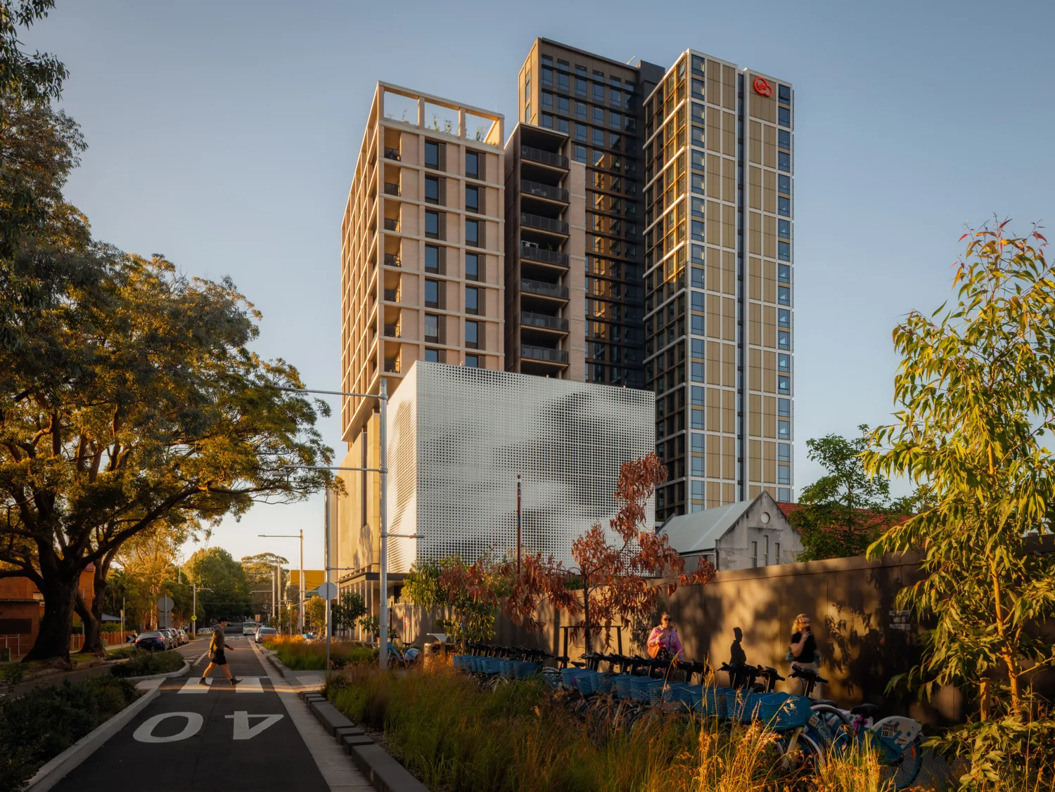 Multi-storey residential tower with balconies above a podium, viewed from a tree-lined street with cycle parking and landscaping.