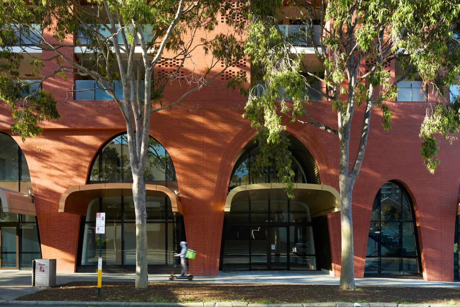 Street-level view of a red brick residential building with large arched entrances, glazed façades, mature trees and a cyclist passing in the foreground.