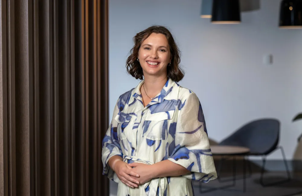 Person standing indoors beside vertical timber wall panels, wearing a patterned light-coloured dress, with seating and pendant lights visible in the background.