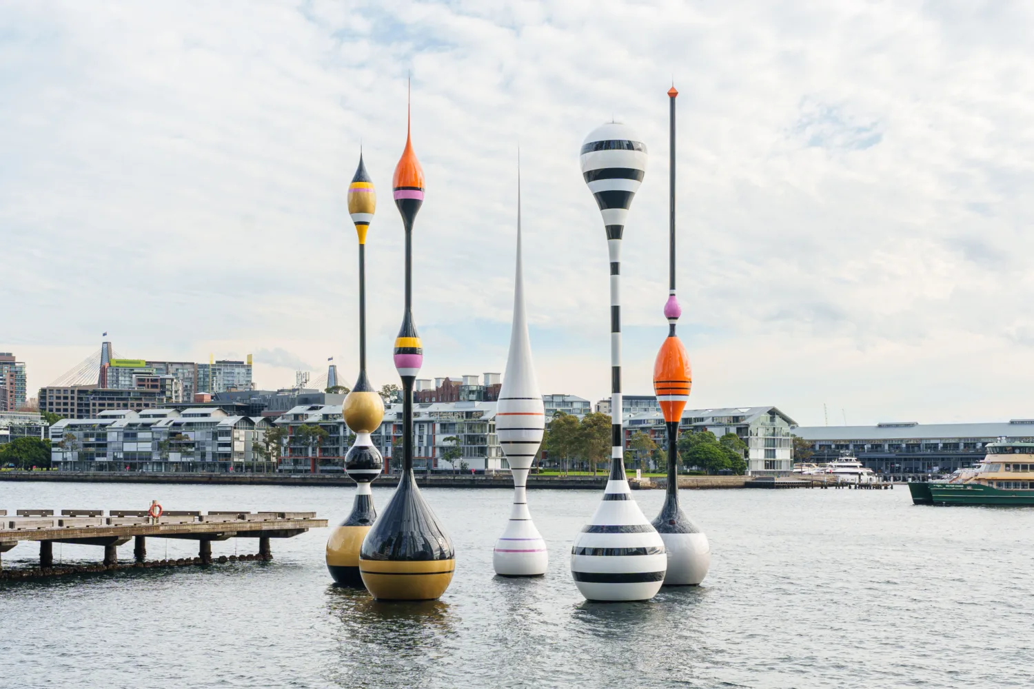 Colourful sculptural buoy-like structures installed in a harbour with waterfront buildings, boats and a pedestrian pier in the background.
