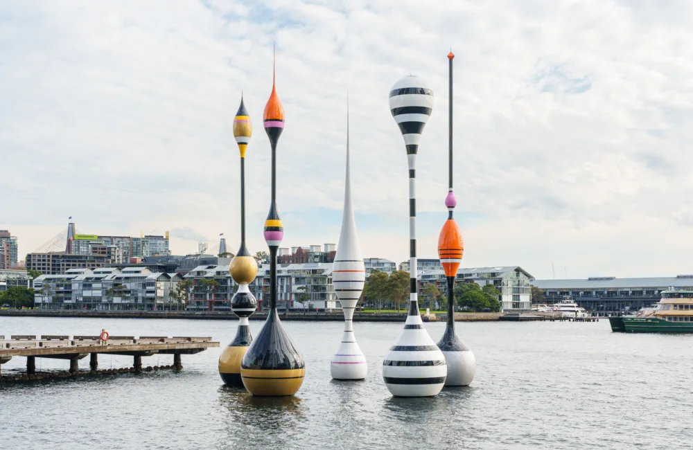 Colourful sculptural buoy-like structures installed in a harbour with waterfront buildings, boats and a pedestrian pier in the background.