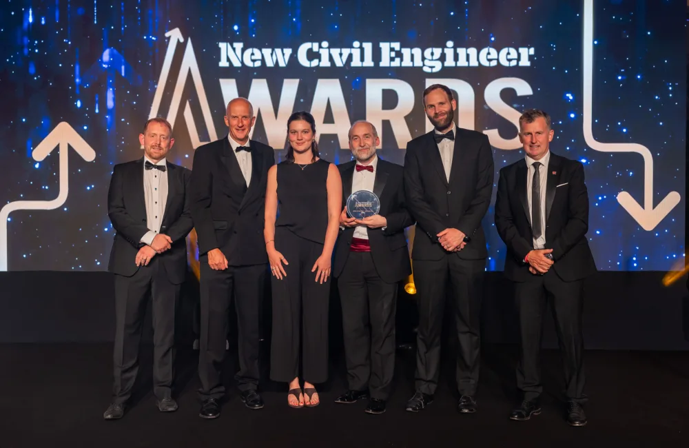 People in formal attire pose on stage at the New Civil Engineer Awards, with one person holding a glass award trophy in front of an illuminated event backdrop.