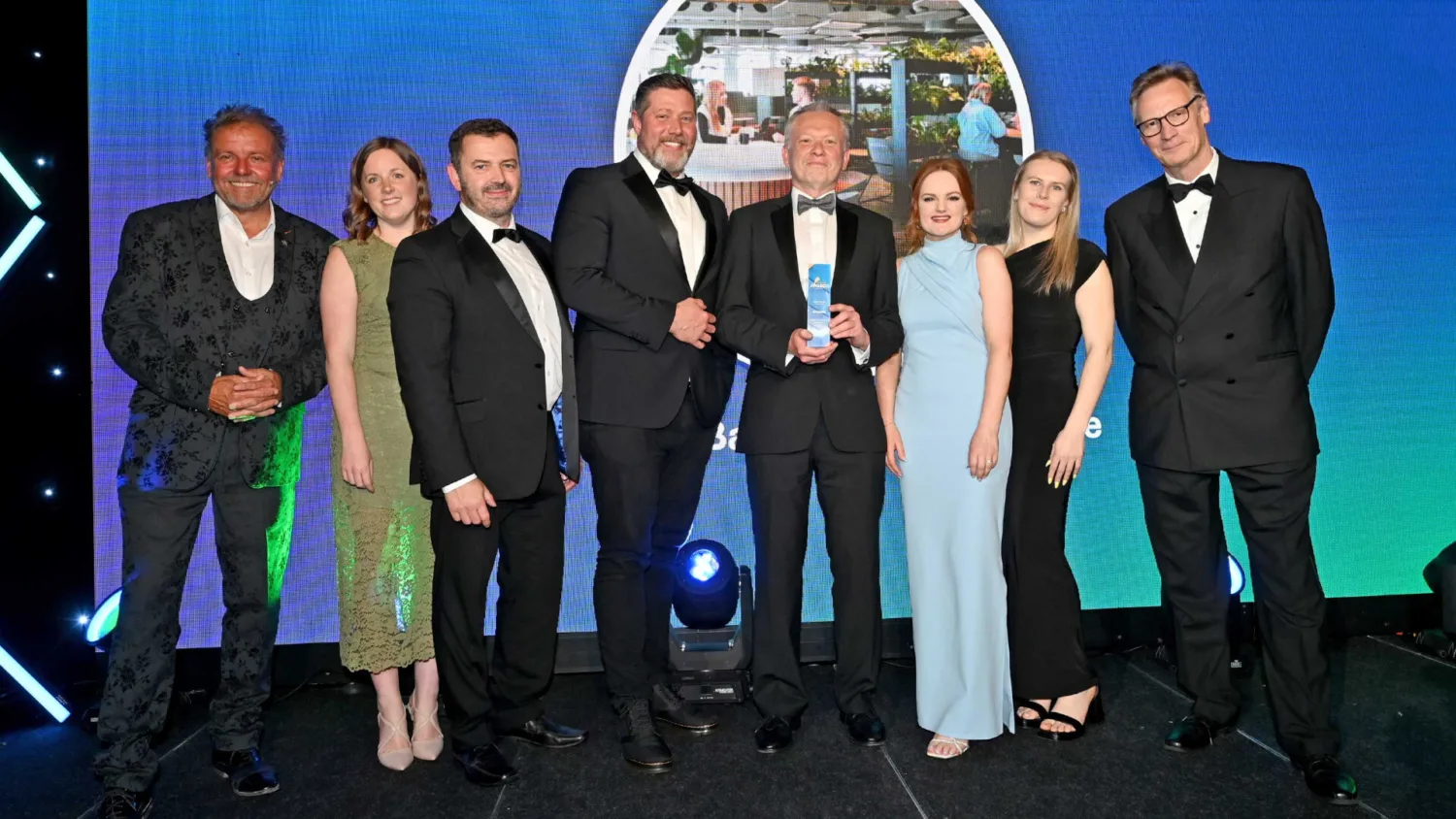 Eight people stand on a stage in formal black and evening attire. Two individuals at center hold a blue glass award. Behind them, a large screen shows an office scene with plants and desks, with stage lighting visible below.