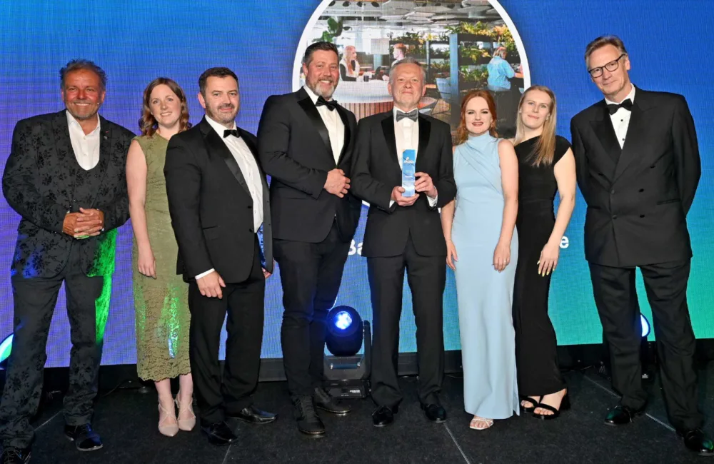 Eight people stand on a stage in formal black and evening attire. Two individuals at center hold a blue glass award. Behind them, a large screen shows an office scene with plants and desks, with stage lighting visible below.