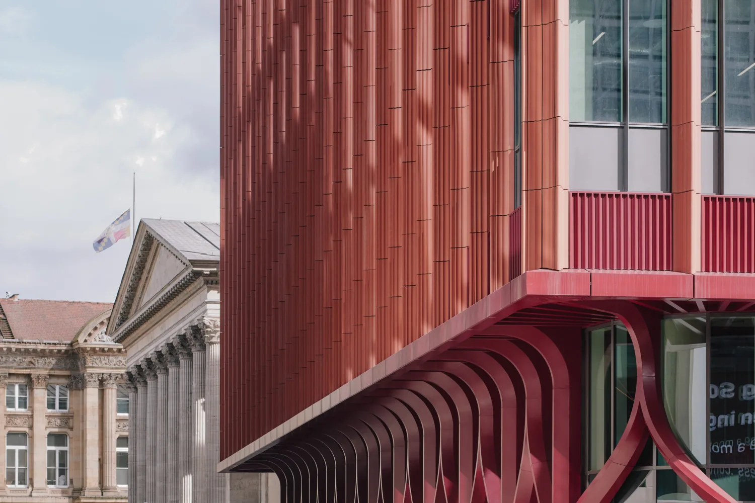 Red modern building with vertical ribbed façade and curved red supports beneath an overhang, large glass windows at right. A classical stone building with columns and a flag appears in the background under a cloudy sky.