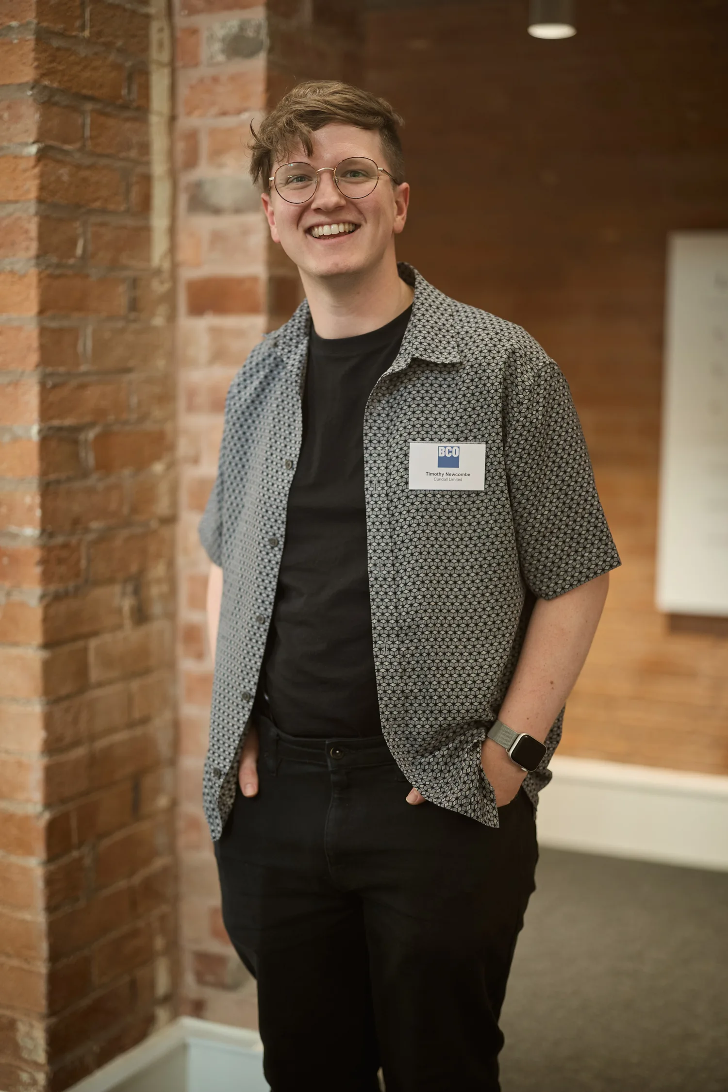 Timothy standing indoors against a brick wall, wearing a patterned short-sleeve shirt over a black t-shirt and black trousers. A name badge with text “BCO” and “Hello my name is” is pinned to the shirt.
